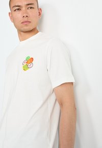 White cotton T-shirt with a round neckline and short sleeves. Features an embroidered design of colorful circles on the chest.