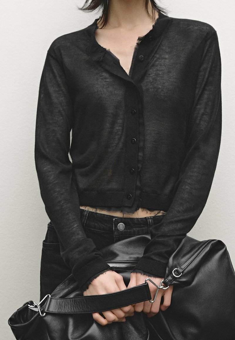Personne portant un cardigan noir boutonné avec des bords effilochés, un pantalon noir, et tenant un sac en cuir noir avec les deux mains.