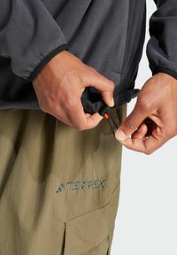 Pullover en polaire de couleur grise avec des poignets noirs, doté de cordons de serrage ajustables. Pantalons vert olive avec le logo Terrex en broderie assortie.