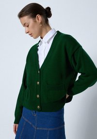 Vrouw met donker haar in een bun, gekleed in gouden oorbellen, een groene vest met knopen, een witte blouse en een blauwe denimrok tegen een effen achtergrond.