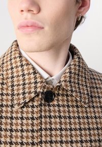 Junge Person mit heller Haut, die einen silbernen Creolenohrring und einen braun-beigen Hahnentrittmantel trägt, der über einem weißen Hemd mit Kragen geschlossen ist.