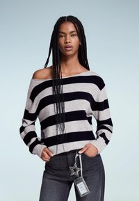 Hosszú fonott hajú nő fekete és szürke csíkos, vállnélküli pulóverben és sötét farmernadrágban, csillag alakú kulcstartó kiegészítővel.