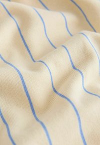 Světlé béžové tkanina s modrými pruhy, s jemnou a hladkou texturou, která se jemně skládá a ukazuje vzor a barevný kontrast.