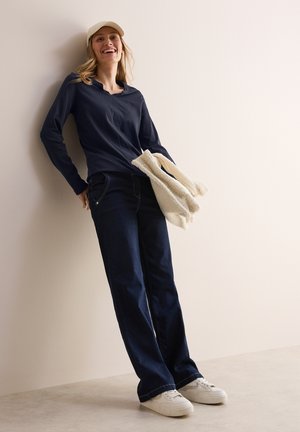 Chemise à manches longues marine, jeans en denim foncé, baskets blanches, tenant un pull léger et moelleux. Design simple, coupe décontractée.