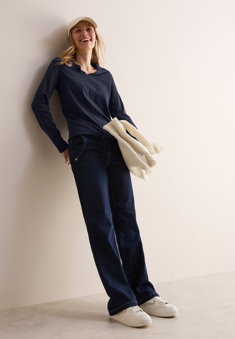 Marineblaues Langarmhemd, dunkle Jeans, weiße Sneakers, hält einen hellen, flauschigen Pullover. Einfaches Design, lockere Passform.