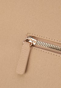 Beige textureret stof med en lille lynlåsdetalje, der har en guldfarvet metaltræk og et tydeligt lynlåsbånd.