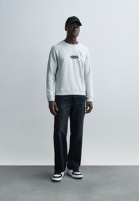 Γκρι φούτερ με μικρό μαύρο λογότυπο, σκούρο τζιν παντελόνι και μαύρο καπέλο· συνδυασμένο με μαύρα και λευκά αθλητικά παπούτσια. Το μοντέλο στέκεται σε γκρι πάτωμα.