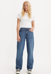 Biała koszulka z krótkim rękawem w zestawieniu z luźnymi, wysokimi dżinsami w niebieskim kolorze. Dżinsy mają klasyczny pięcio-kieszeniowy krój i lekkie przetarcia.