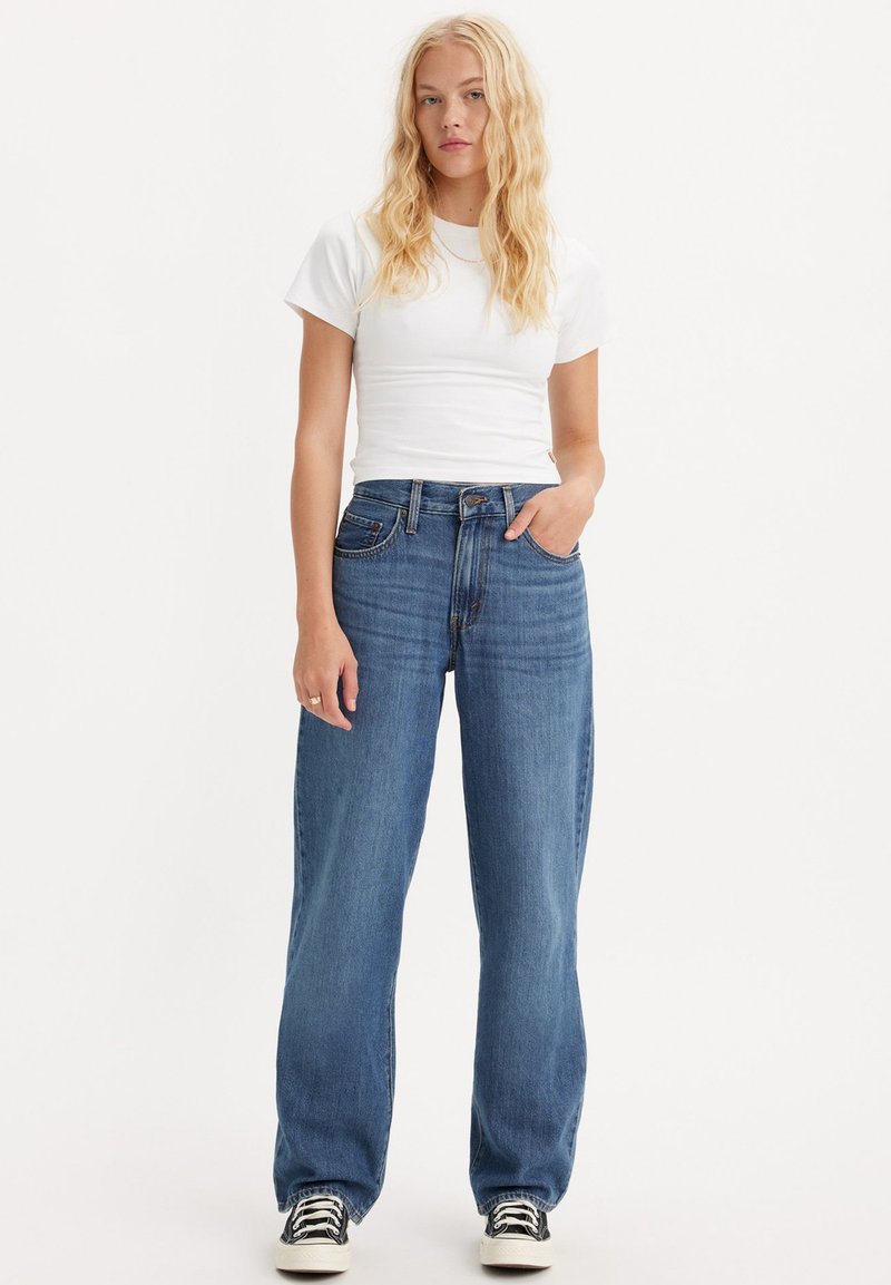 Biała koszulka z krótkim rękawem w zestawieniu z luźnymi, wysokimi dżinsami w niebieskim kolorze. Dżinsy mają klasyczny pięcio-kieszeniowy krój i lekkie przetarcia.