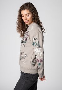 Young woman with curly hair wearing beige sweatshirt with colorful doodles and words, paired with dark pants, against plain background.