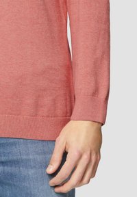 Koraalrood sweater gemaakt van zachte stof, met ribgebreide manchetten en een relaxte pasvorm, gecombineerd met lichtblauwe denim.