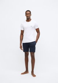 Bílé bavlněné tričko s malým černým logem "BOSS" na hrudi, doplněné tmavě modrými šortkami se stejným logem, model stojí bosý.