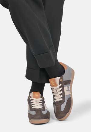 Person iført brune, beige og grå snøresneakers med beige snørebånd og sorte bukser, stående med krydsede fødder.