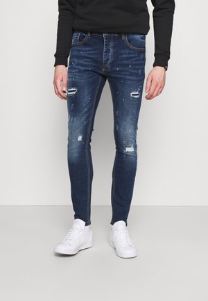 Jeans in denim blu a vestibilità slim, con dettagli strappati, leggera scoloritura e toppe sfrangiate, abbinati a sneaker bianche su uno sfondo neutro.