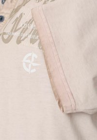 Beige Baumwoll-T-Shirt mit Kragen, ausgestattet mit Knöpfen, einer glatten Textur und bedruckten Texten sowie Logoakzenten in Hellgrau und Weiß.