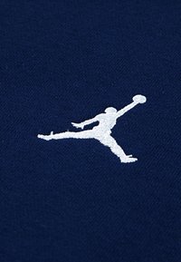 Námornícka látka s hladkým povrchom, na ktorej sa nachádza biely vyšitý logotyp Jumpman, znázorňujúci postavu skákajúcu s basketbalom.
