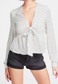 Blouse blanche à pois noirs, col en V, devant noué, manches longues et ourlet fluide ; associée à un short en denim noir.