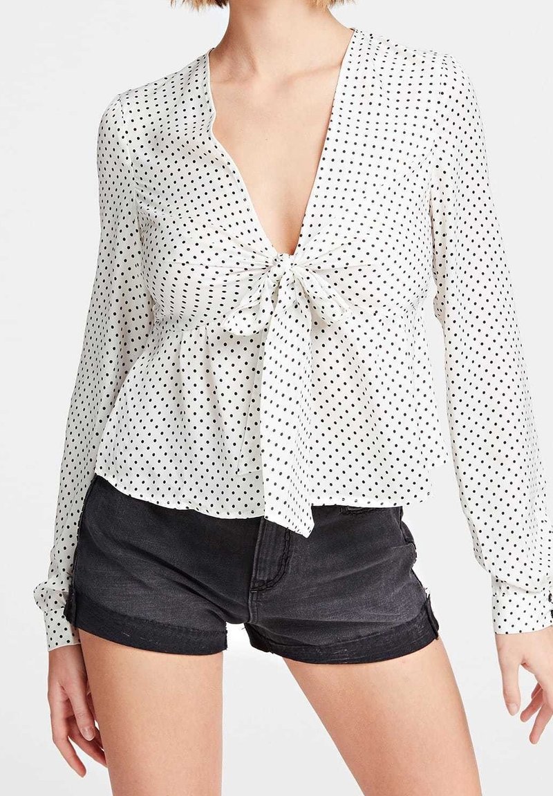 Blouse blanche à pois noirs, col en V, devant noué, manches longues et ourlet fluide ; associée à un short en denim noir.