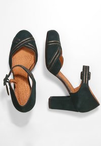 Chaussures à talons hauts en suede vert foncé avec des brides croisées, des accents dorés et une doublure beige. Talon carré et détail de bride de cheville.