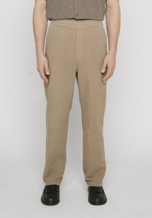 Mann iført beige bukser med rette ben og svarte skinnloafers, stående mot en enkel lys bakgrunn.