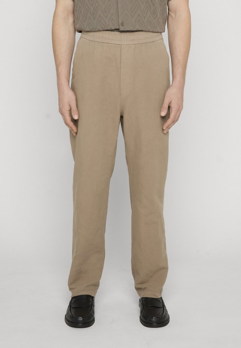 Homme portant un pantalon beige à jambe droite et des mocassins en cuir noir, debout contre un fond clair uni.
