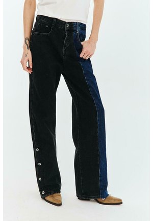 Osoba ubrana w czarne i niebieskie dżinsy o szerokich nogawkach z guzikami na dolnym, zewnętrznym szwie oraz jasnobrązowe, spiczaste buty, stojąca z jedną ręką w kieszeni.