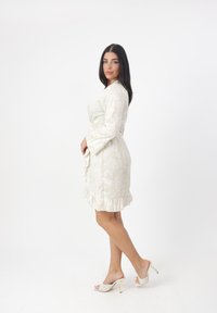 Helles beigefarbenes Blumenkleid mit langen Ärmeln, gerafftem Saum und gebundenem Taillengurt. Kombiniert mit weißen offenen Sandalen mit einem gemusterten Design.