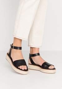 Fötter i svarta ankelsandaler med kilklack och beige vävda plattformssulor, iförda tillsammans med avkortade off-white byxor mot en neutral bakgrund.