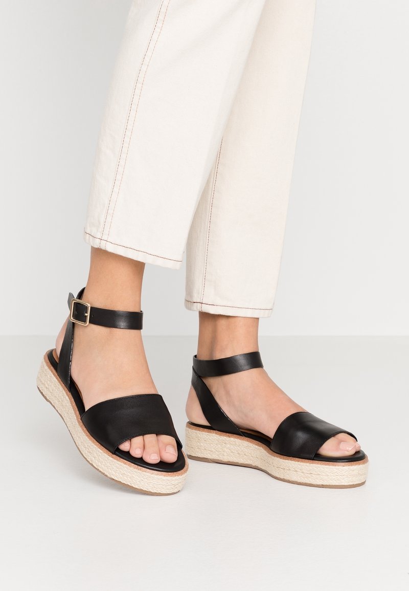 Fötter i svarta ankelsandaler med kilklack och beige vävda plattformssulor, iförda tillsammans med avkortade off-white byxor mot en neutral bakgrund.