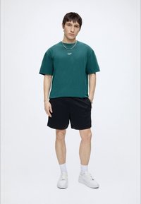 Jeune homme debout portant une chemise verte à manches courtes, un short noir, des chaussettes blanches et des baskets blanches sur un fond uni.