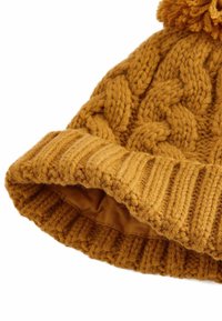 Stickad senapsfärgad mössa med ett flätmönster, ribbad manschett och en fluffig pom-pom på toppen; mjuk textur och rundad form.