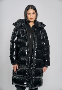 Czarna pikowana kurtka puchowa o błyszczącym wykończeniu, z kapturem i pikowaną fakturą. Posiada zamek błyskawiczny i boczne kieszenie, noszona na ciemnym swetrze.