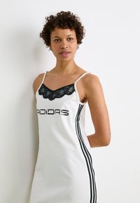 Bela brezrokavna obleka z črnimi čipkastimi robovi, ki vključuje logotip Adidas in črne vertikalne črte po obeh straneh. Mehka tkanina z oprijetim krojem.