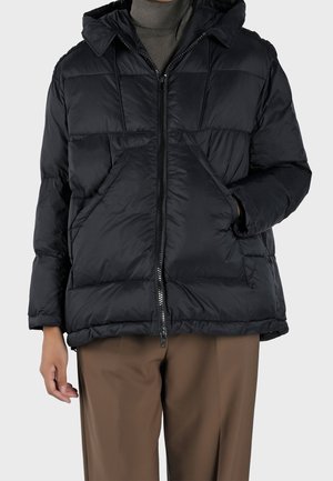Giacca a vento nera con cappuccio, chiusura a zip e ampie tasche frontali. Presenta un design trapuntato e polsini elastici. Indossata sopra pantaloni marroni.