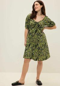 Grünes und schwarzes gemustertes Kleid mit kurzen Puffärmeln und V-Ausschnitt, das mit einem gerafften Vorderteil versehen ist. Getragen mit schwarzen Slippers.