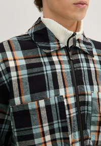 Kariertes Jackett mit einer Mischung aus Schwarz, Blau und Orange, einem hohen weißen Rippkragen und einem frontalen Reißverschluss. Zwei Fronttaschen.