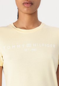 Gul bomull t-skjorte med korte ermer, med en hvit "TOMMY HILFIGER EST. 1985" logo på brystet. Myk tekstur, rund hals.
