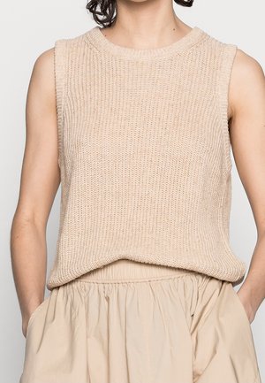 Kvinde iført en ærmeløs beige striktop og en højtaljet beige nederdel, står med hænderne i nederdelens lommer mod en ensfarvet baggrund.