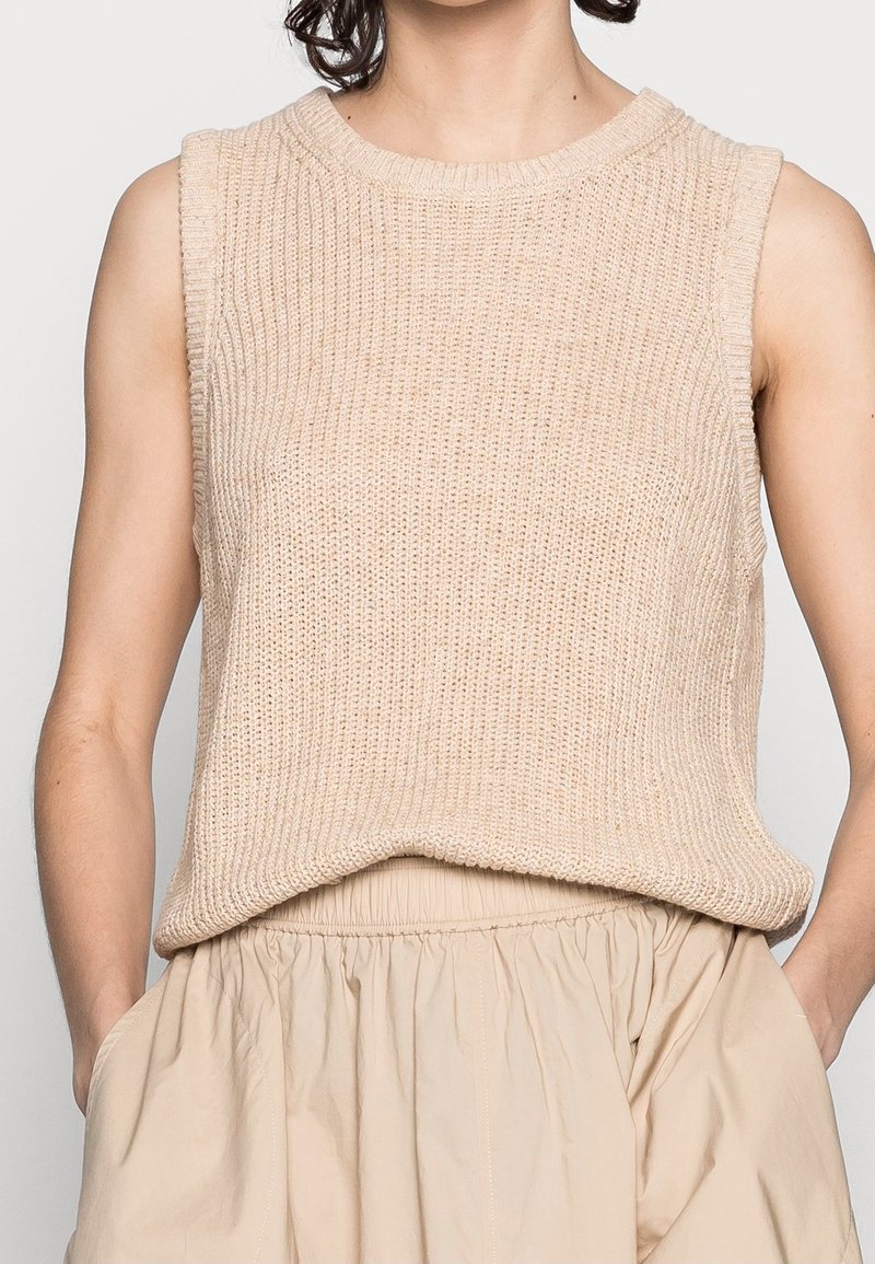 Femme portant un haut en maille beige sans manches et une jupe beige taille haute, debout les mains dans les poches de la jupe devant un fond uni.