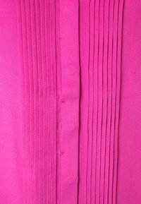 Tissu plissé fuchsia présentant des plis verticaux d'un côté et une texture lisse de l'autre, avec des coutures visibles et des détails de boutons.