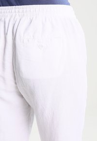 Bílé lněné kalhoty s elastickým pasem a jednou zadní kapsou se zapínáním na knoflík. Měkká textura, volný střih.