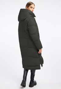 Lang, donkergroen pufferjas met capuchon, gekenmerkt door een gequilt ontwerp en zijzakken. Gemaakt over zwarte broek en forse zwart laarzen.
