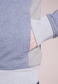 La mano di una persona all'interno della tasca di una felpa color block blu e grigia con polsini e orlo a costine.