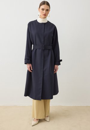 Frau trägt einen dunklen, langen Mantel mit Gürtel über einem weißen Rollkragenpullover und beigen, weiten Hosen, steht vor einer einfachen hellen Wand.
