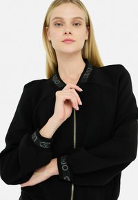 Veste bomber noire avec fermeture éclair à l'avant, épaules rembourrées et poignets à motif texturé. Tissu doux avec un léger éclat.