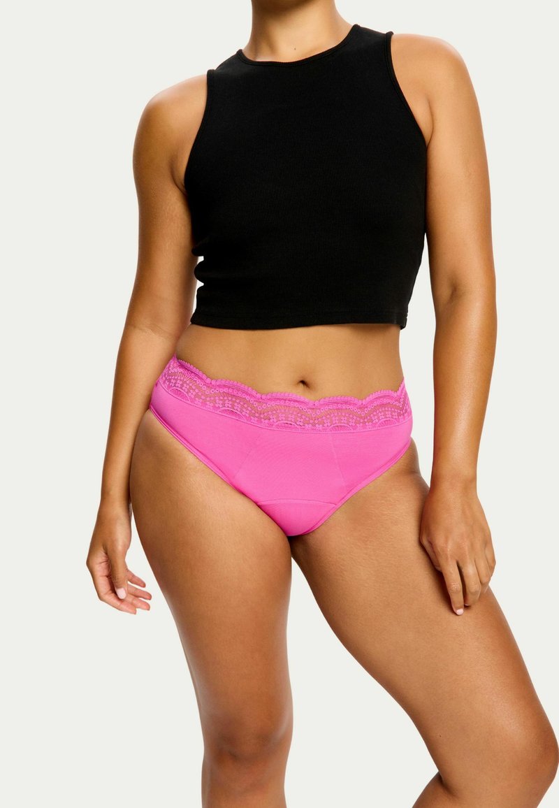 Culotte rose avec une taille en dentelle, fabriquée à partir d'un tissu doux et extensible. Portée avec un haut sans manches noir, mettant en valeur le détail de la taille.