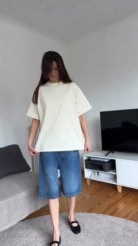 Femme à l'intérieur portant un t-shirt blanc ample, un short en jean mi-long et des ballerines noires, debout sur un tapis rond gris dans un salon.