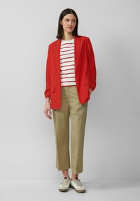 Blazer rosso, camicia a righe bianche e rosse, pantaloni khaki e sneakers casual con accenti bianchi e beige. Texture lisce e forme su misura.