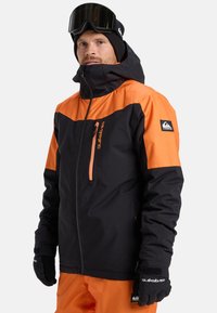 Giacca invernale nera e arancione con tasca con zip, cappuccio regolabile e dettagli con logo. La trama appare liscia e resistente.