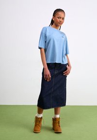 Světlomodré oversized tričko, tmavé deninové midi sukně a béžové kotníkové boty se žlutými šňůrkami. Modelka stojí na zelené podlaze s rukama na bok.